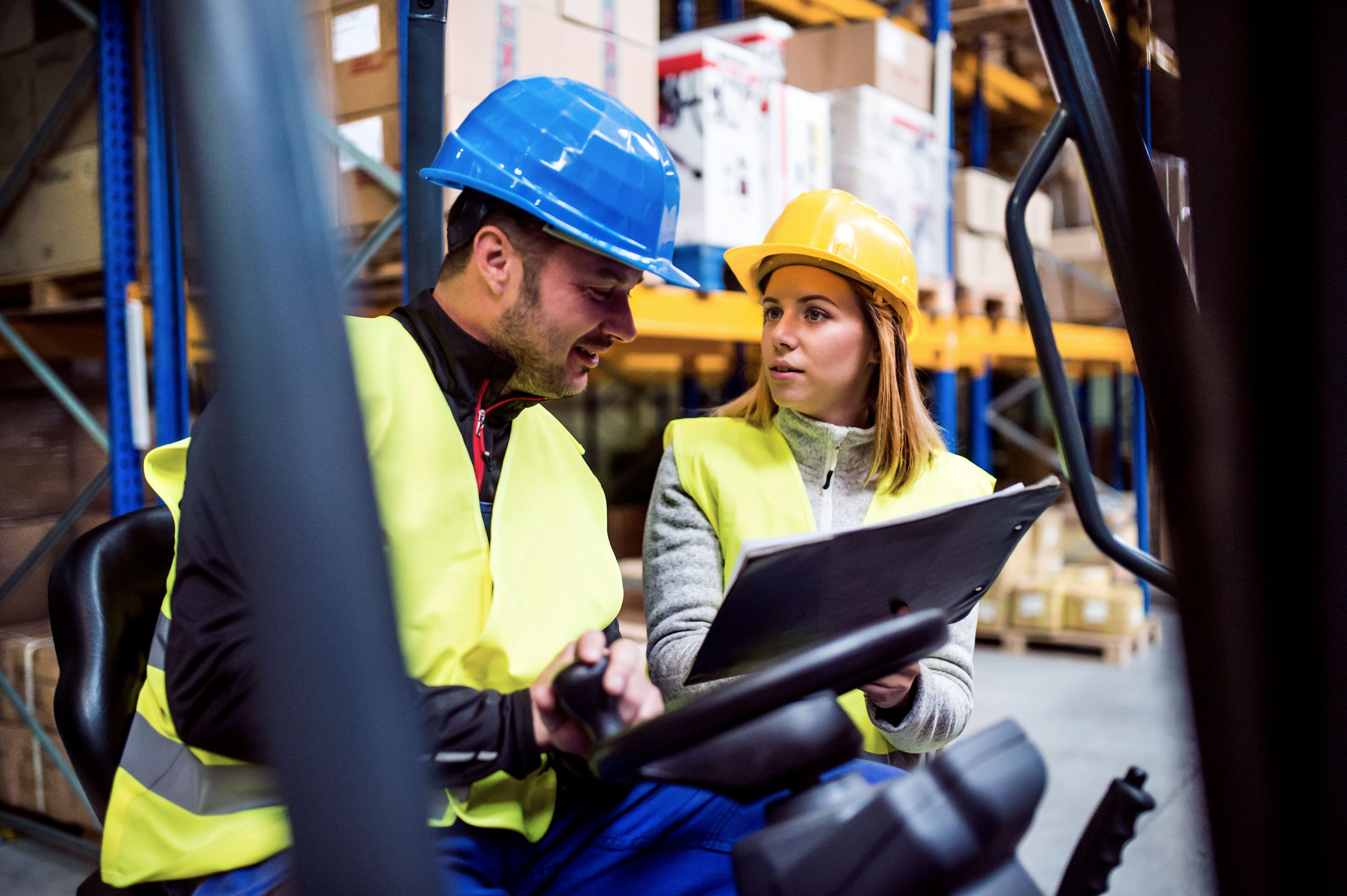 Warehouse workers in safety gear discussing logistics while operating a forklift.