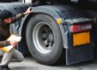 Vehicle inspector examining a lorry's rear tyre with a clipboard during a safety check.