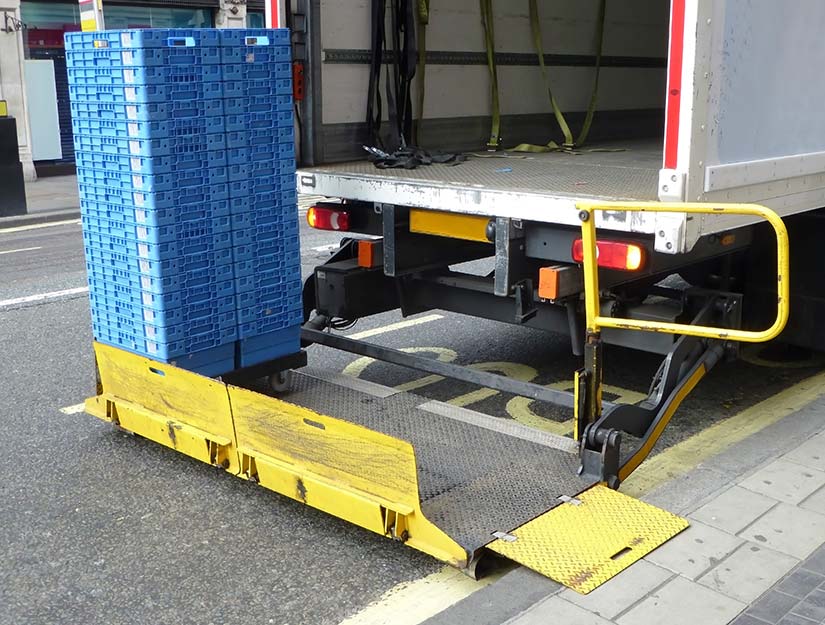 Delivery truck with open tail lift unloading stacked blue crates onto a high street.