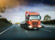 Red articulated lorry driving on a motorway during early evening with headlights on.