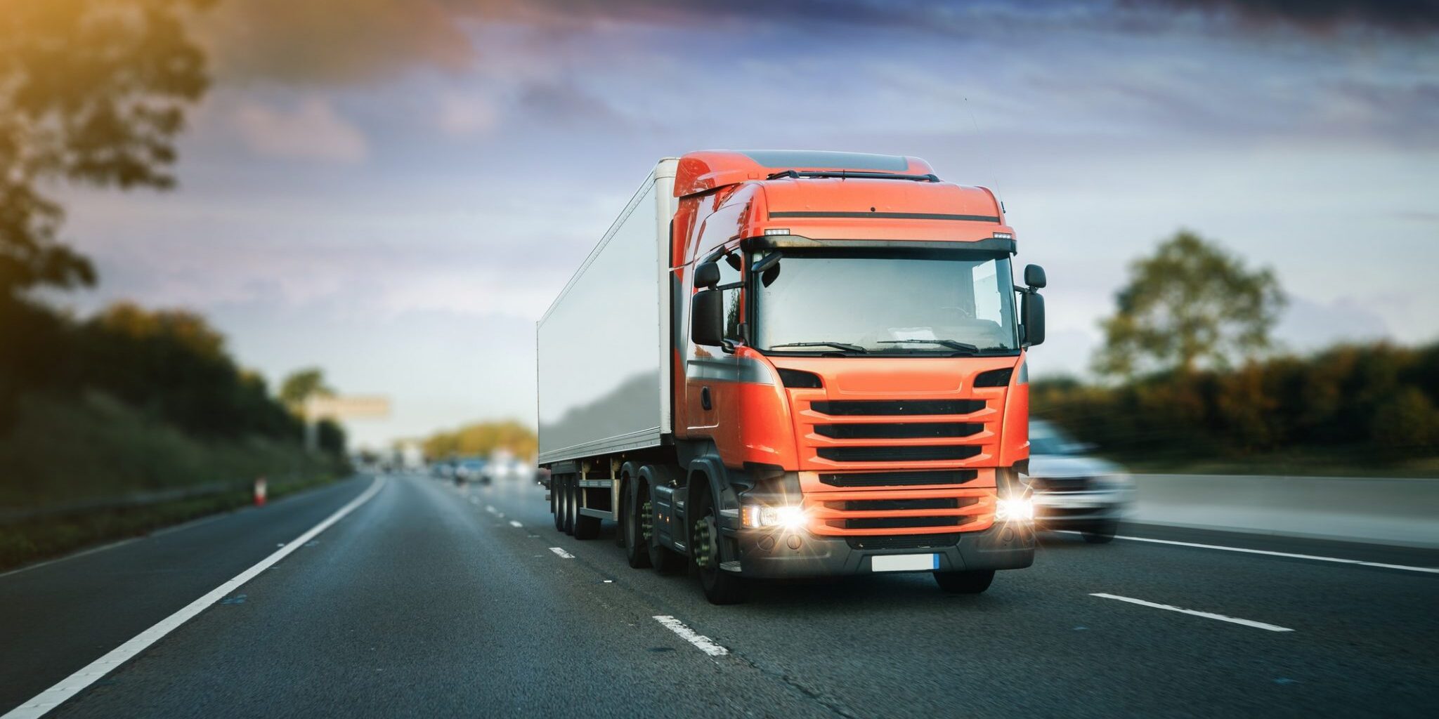 Red articulated lorry driving on a motorway during early evening with headlights on.
