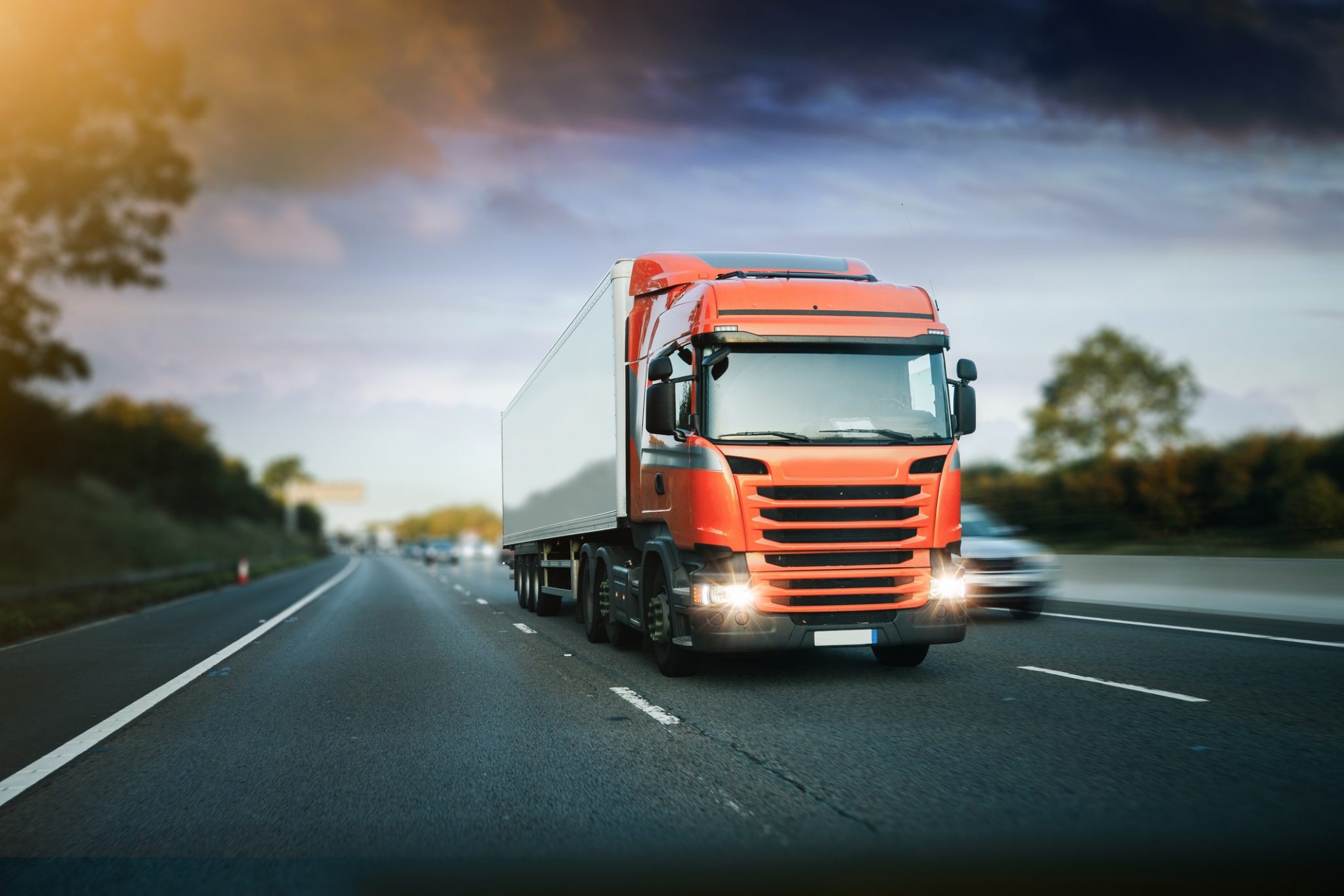 Red articulated lorry driving on a motorway during early evening with headlights on.